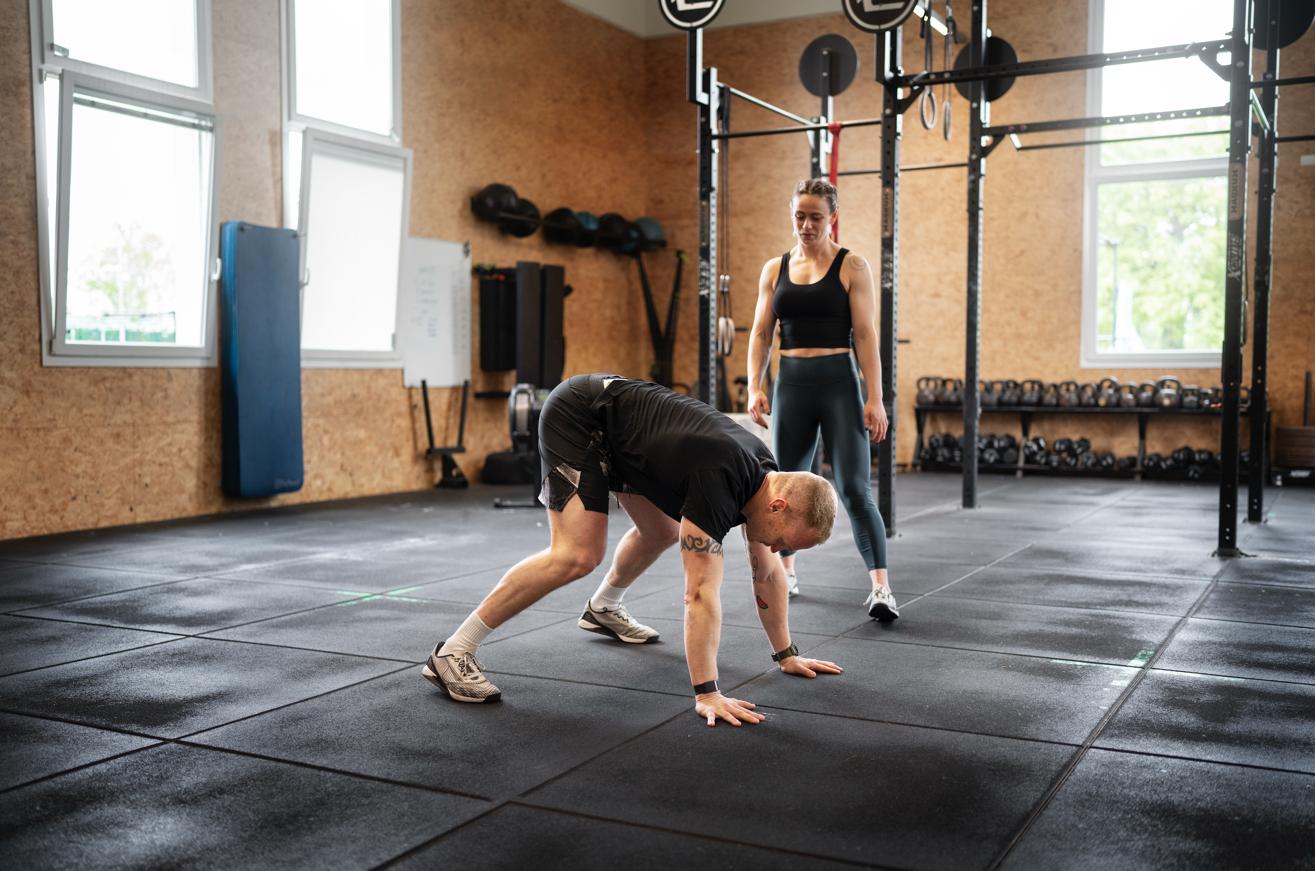 Two people training in a gym with black rubber flooring tiles, showing the padded surface used for functional and strength workouts.
