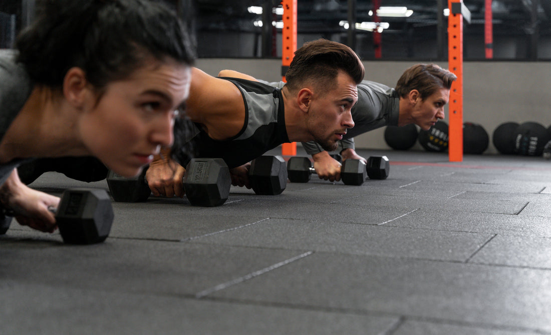 A group of athletes performs dumbbell push-ups on interlocking rubber flooring in a gym setting.